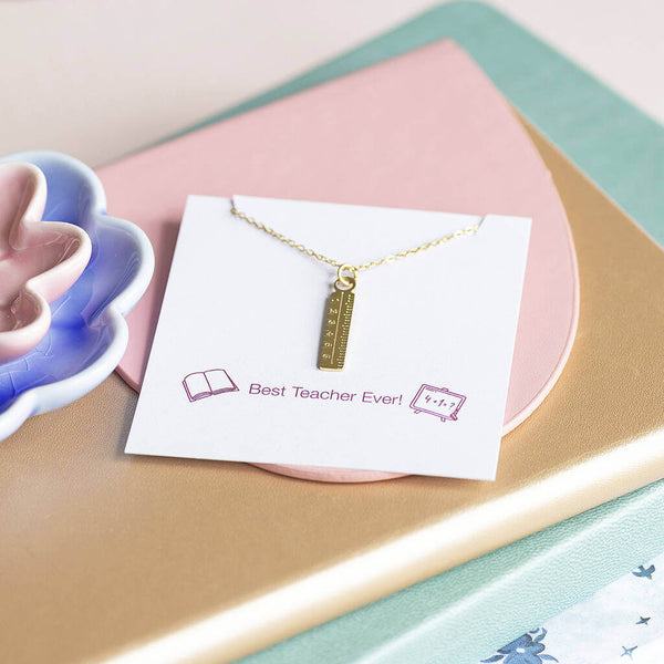 A gold plated dainty ruler charm necklace presented on a Best Teacher Ever! sentiment card sitting on a pink and gold leather Teacher's notebook.