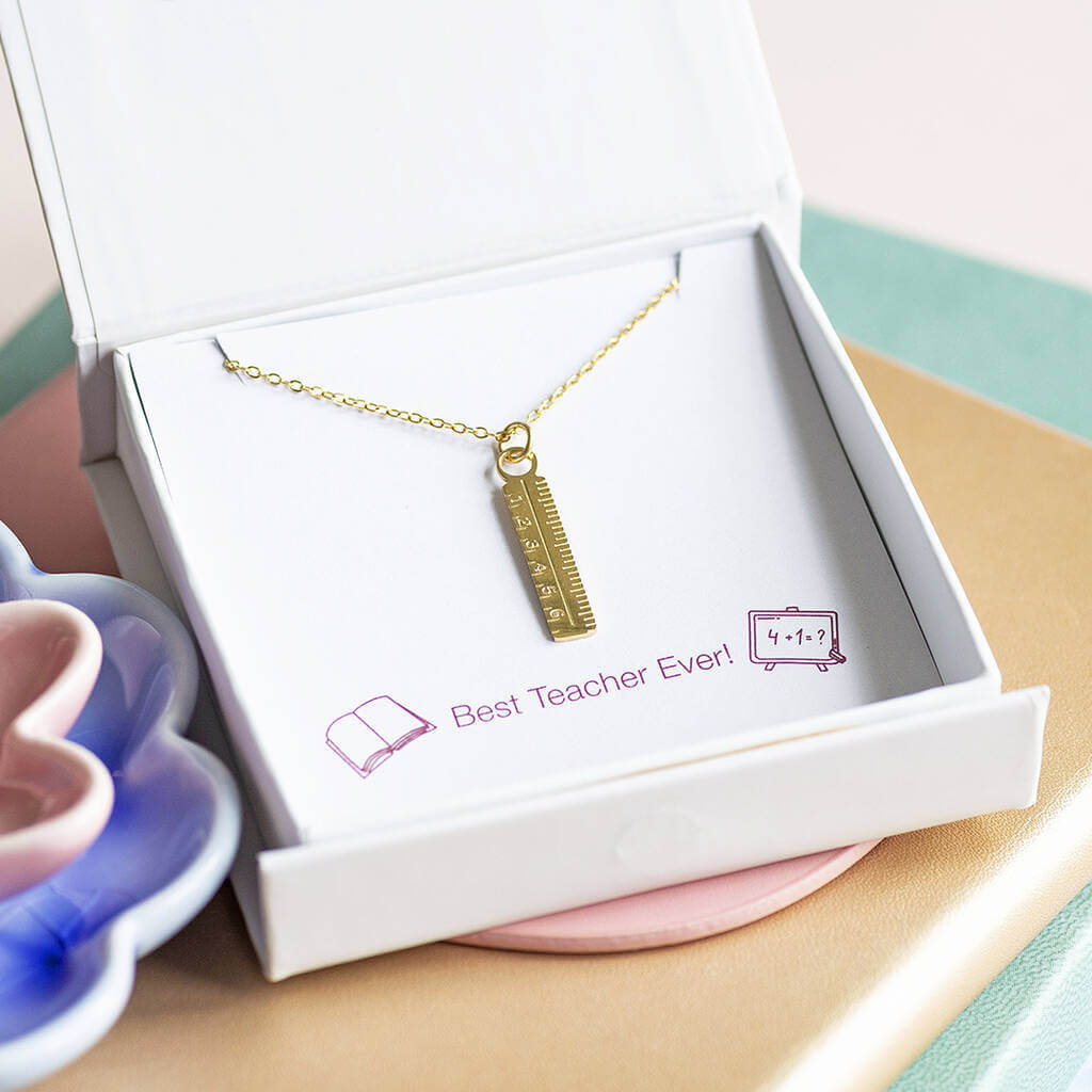 A gold plated dainty ruler charm necklace presented on a Best Teacher Ever! sentiment card inside a Joy by Corrine Smith gift box. Jewellery dishes are in the background.