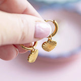 Gold plated heart hoop earrings with a sunburst design on the heart being held by a pair of women's hands.