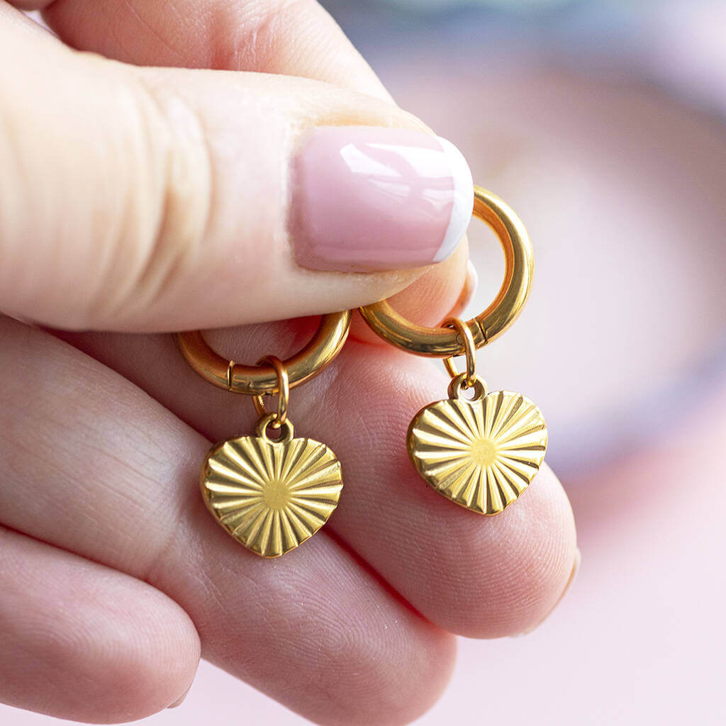 Gold plated heart hoop earrings with a sunburst design on the heart displayed on a woman's hand.