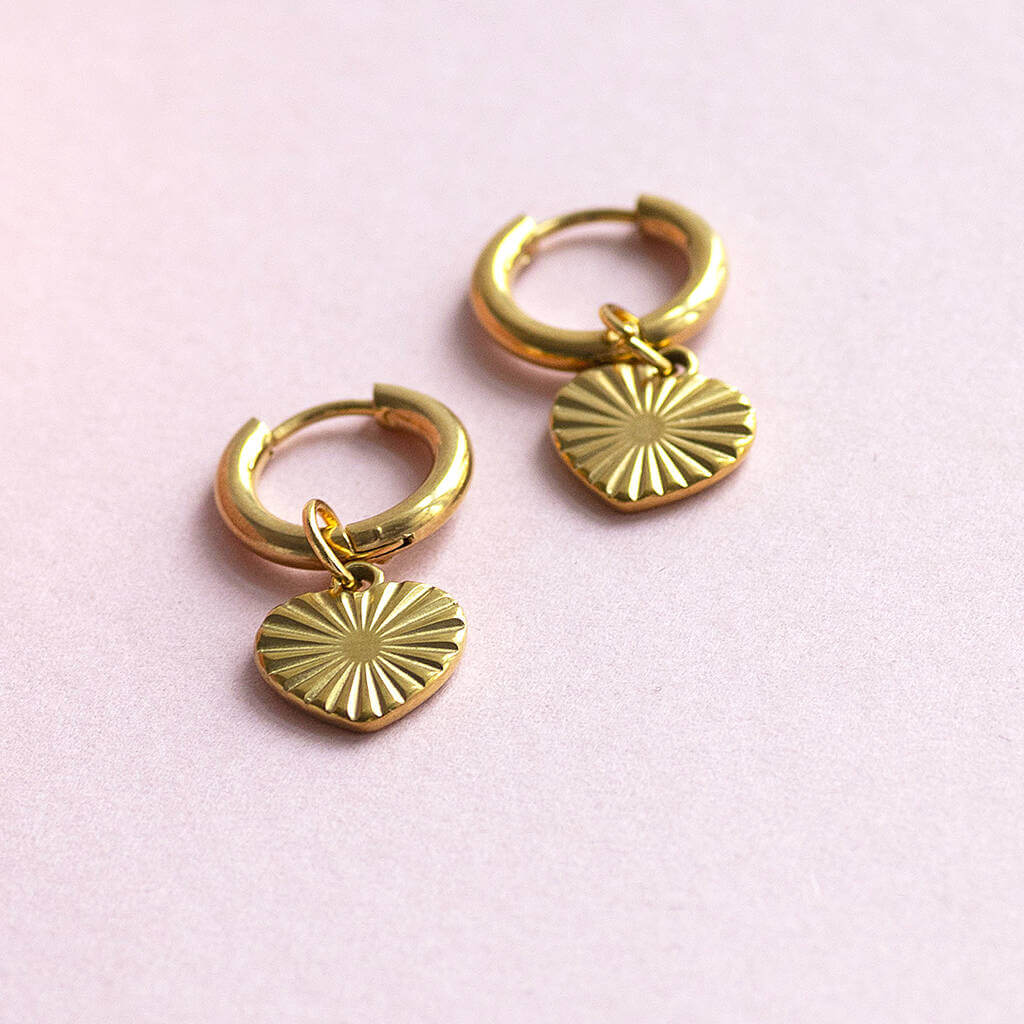 Gold plated heart hoop earrings with a sunburst design on the heart displayed on a pink background.