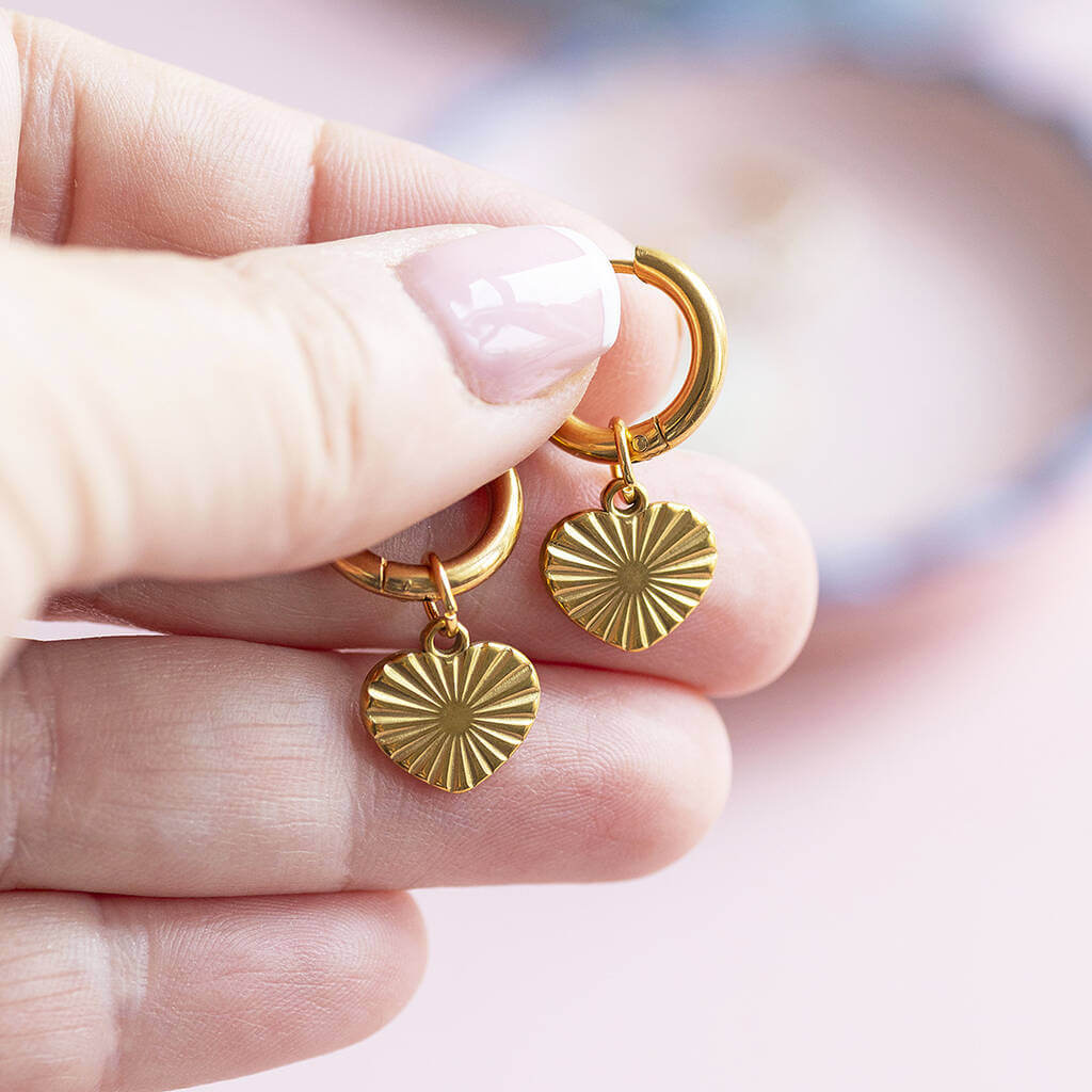 Gold plated heart hoop earrings with a sunburst design on the heart being held by a pair of fingers.