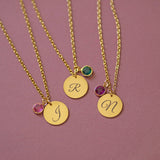 Three gold script initial necklaces with colourful birthstone charms, layered jewellery styling on pink backdrop