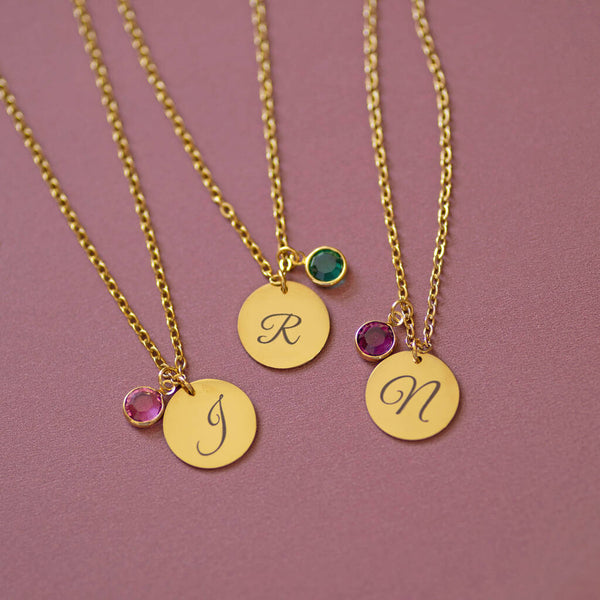 Three gold script initial necklaces with colourful birthstone charms, layered jewellery styling on pink backdrop
