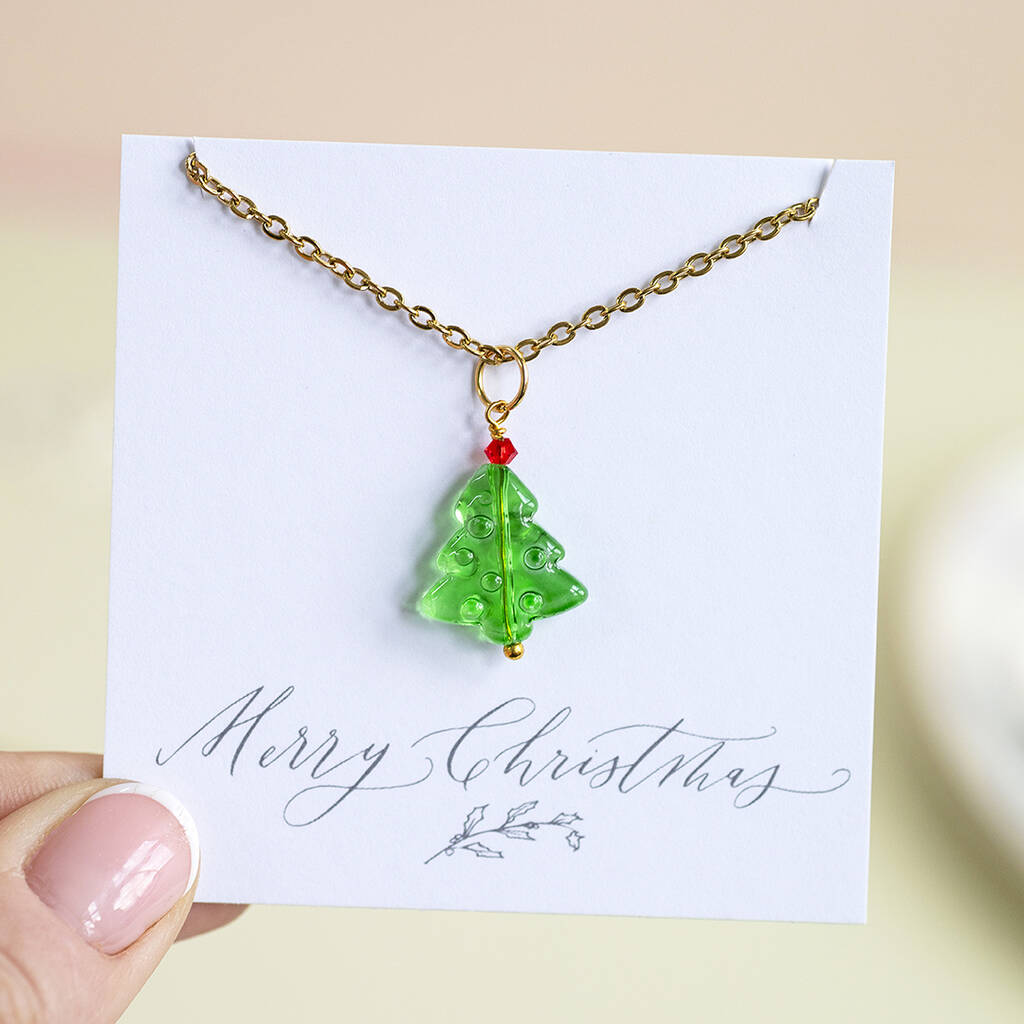 Image shows a green glass Christmas tree pendant necklaces with a red bed bauble at the top of the tree. The pendant is on a gold chain and presented on a Merry Christmas sentiment card.