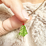 Image shows a green glass Christmas tree pendant necklaces with a red bed bauble at the top of the tree. The pendant is on a gold chain and is worn around the neck with a beige fur jacket.