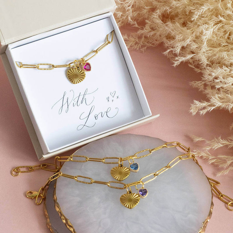 Image shows three gold plated heart charm bracelet with birthstone heart on a pink backdrop. From top to bottom: January birthstone in a gift box with a "with love" sentiment card, March birthstone and February birthstone.