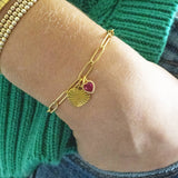 Images shows model in green cardigan and jeans wearing a gold plated heart charm bracelet with Ruby July Birthstone heart charm