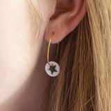 Image shows gold hoop earring with a Mother of Pearl round disc featuring a gold star. Worn on an earlobe.