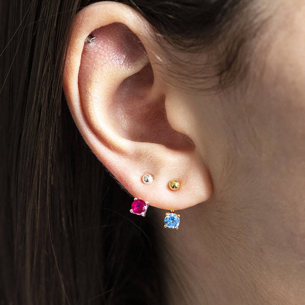 Two pairs of Illusion Birthstone Stud Earrings with a classic round-cut gemstone set in a minimalist prong setting, designed to showcase the birthstone's vibrant colour and clarity. One silver October birthstone earring and one gold December birthstone worn on an earlobe.