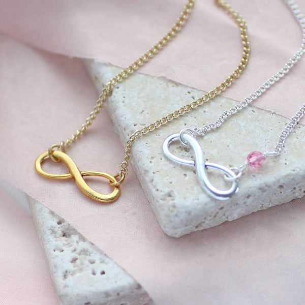 image shows silver infinity birthstone necklace with a Rose crystal and a gold infinity birthstone necklace presented on a piece of peach silk material