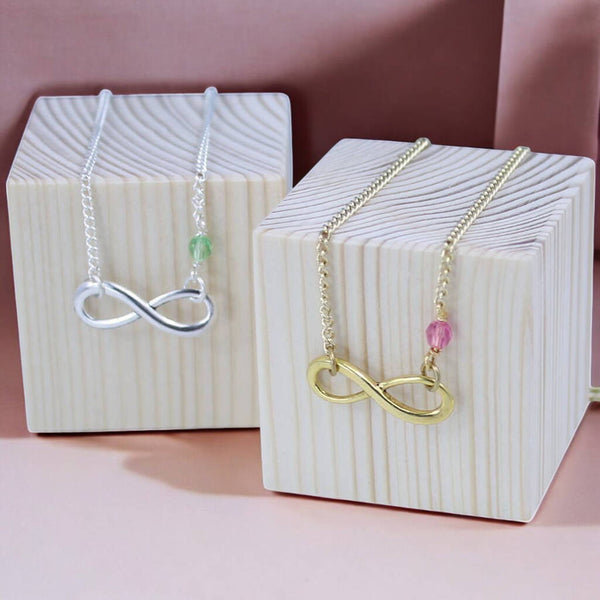 image shows silver infinity birthstone necklace with a Peridot crystal and a gold infinity birthstone necklace with a Rose birthstone presented on wooden jewellery blocks