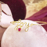 Image shows Gold Plated JOY Script Bracelet with January Birthstone Heart Detail on a pink backdrop.