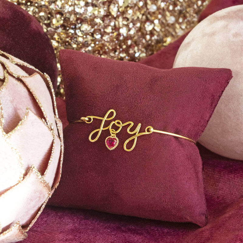 Image shows Gold Plated JOY Script Bracelet with January Birthstone Heart Detail on a maroon jewellery pillow