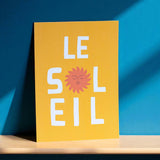 Yellow French wall art print featuring Le Soleil text and sunshine design display against a blue wall