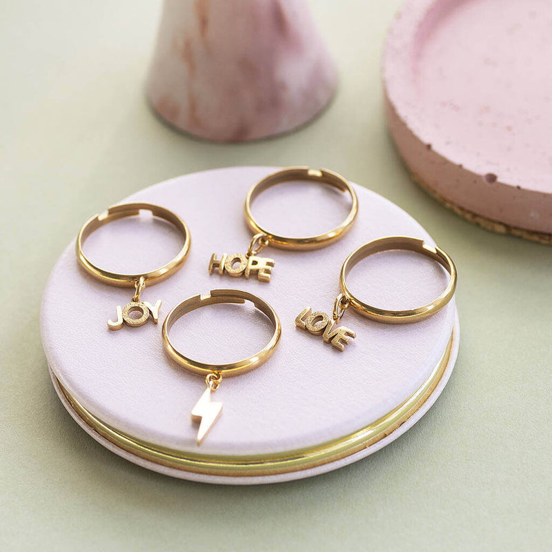 Love, Joy, hope and lighting bolt gold plated adjustable charm rings set on a pink ceramic jewellery trinket plate.