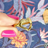 Image shows hand holding love sign gold plated ring in front of a colourful background