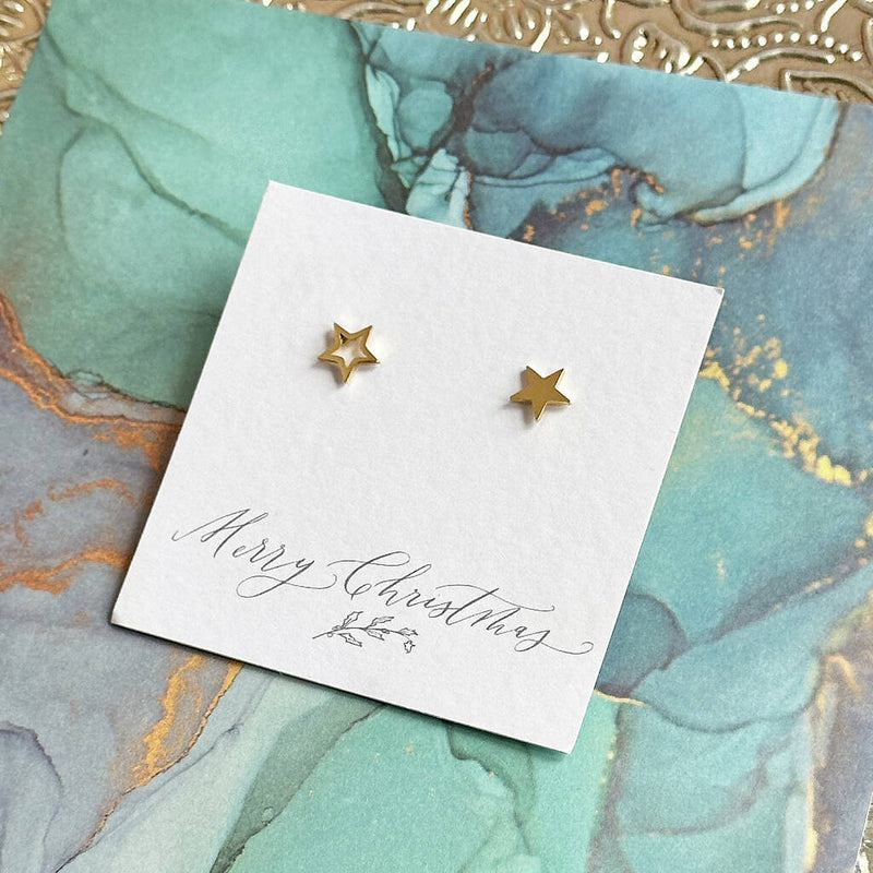 Image shows a pair of gold star stud earrings on a merry christmas sentiment card