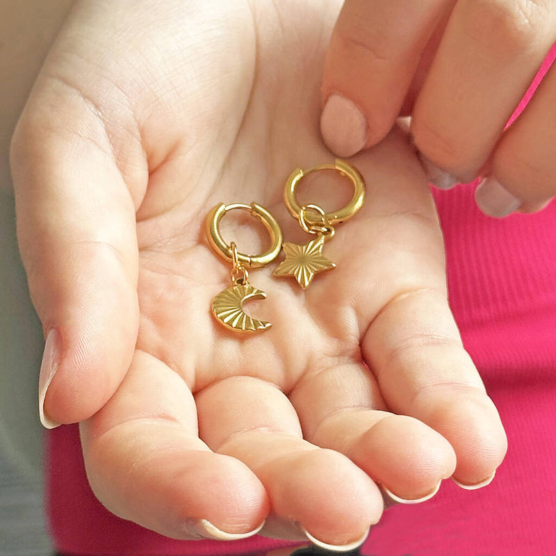 Image shows a hand holding a pair of gold moon and star hoop earrings