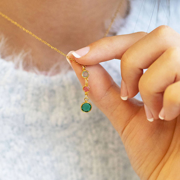 Gold birthstone drop necklace with neutral, pink and green crystal charms held on a blue knit outfit
