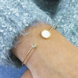 Gold torque bangle worn on a wrist. Banlge feature a round pearl disc and and at the opposite end a smaller crystal disc