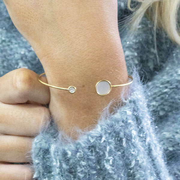 Gold torque bangle with a mother of pearl large disc and crystal small disc at either end of the bangle. Worn on a wrist with a blue fluffy jumper in the background.