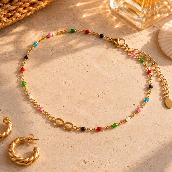 Gold infinity anklet with vibrant multicoloured beads styled with matching gold jewellery on a sunlit background