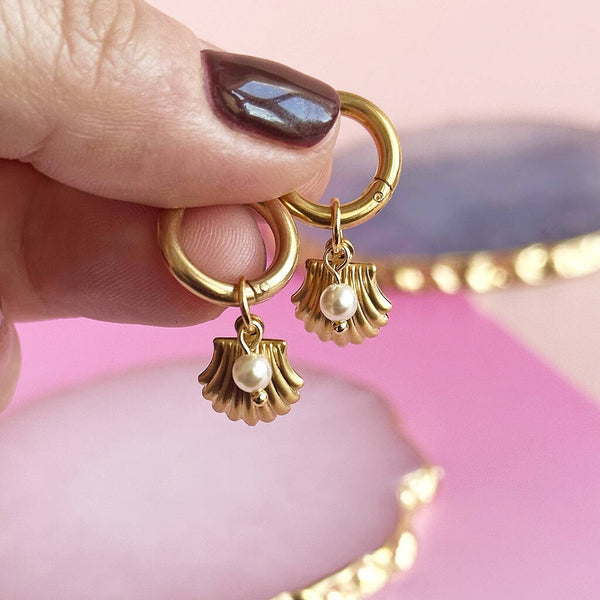images shows a person's hand holding up a pair of gold plated hoop earrings with Oyster shell and pearl charm detail