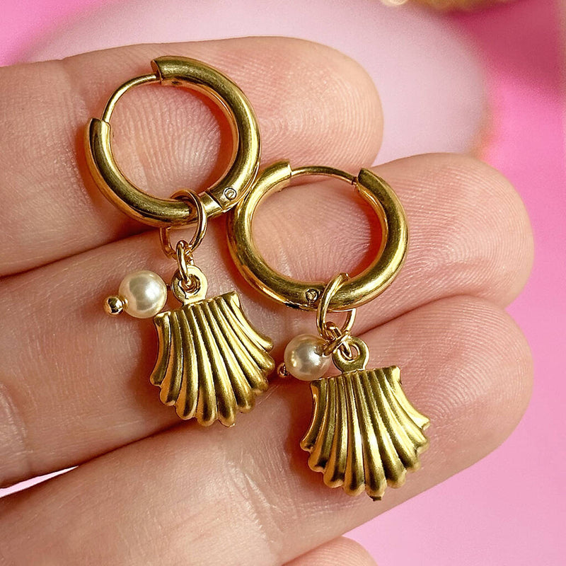 Image shows fingers holding gold plated oyster shell earrings with peark detail in front of a pink background