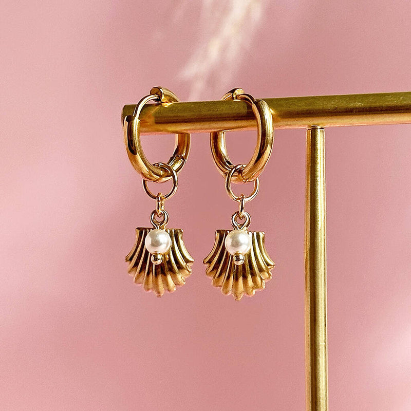 Image shows oyster shell and pearl earrings hanging in front of a pink backgroud
