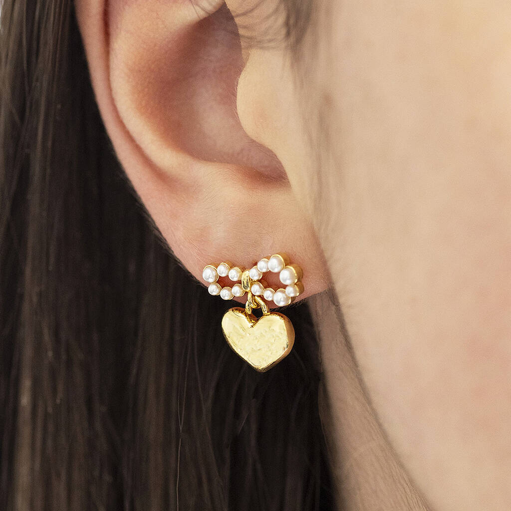 A person wearing an elegant gold earring featuring heart-shaped charm suspended from a dainty pearl bow.