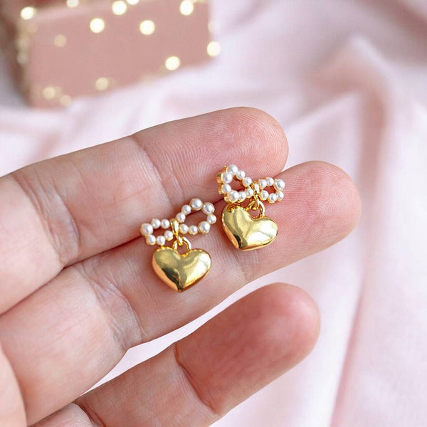 A pair of elegant gold earrings featuring heart-shaped charm suspended from a dainty pearl bow. The earrings are being held in a hand with a pink backdrop.