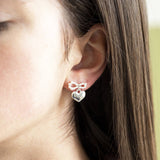 A person wearing an elegant silver earring featuring heart-shaped charm suspended from a dainty pearl bow.