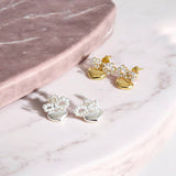 Two pairs of elegant earrings, one silver pair and one gold pair, featuring heart-shaped charms suspended from small pearl bows displayed on a pink marble dressing table top.