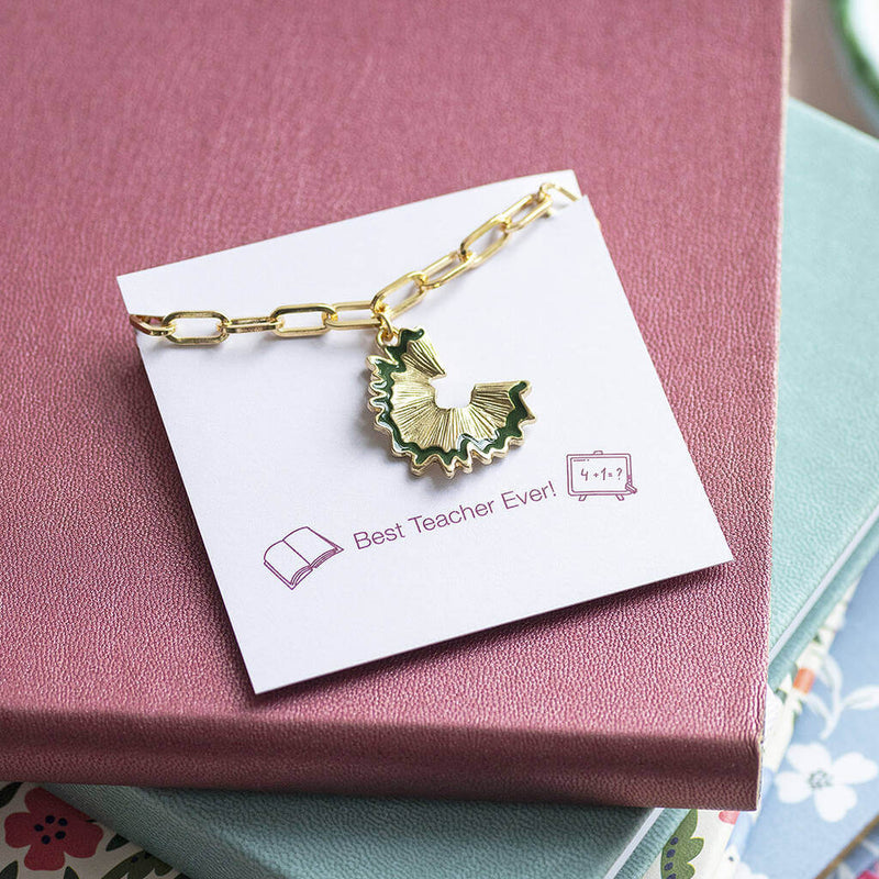 Gold plated charm bracelet with a hanging gold with green trim pencil sharping charm, presented on a Best Teacher Ever sentiment card. Placed on top of a pink , blue and floral notebook stack.