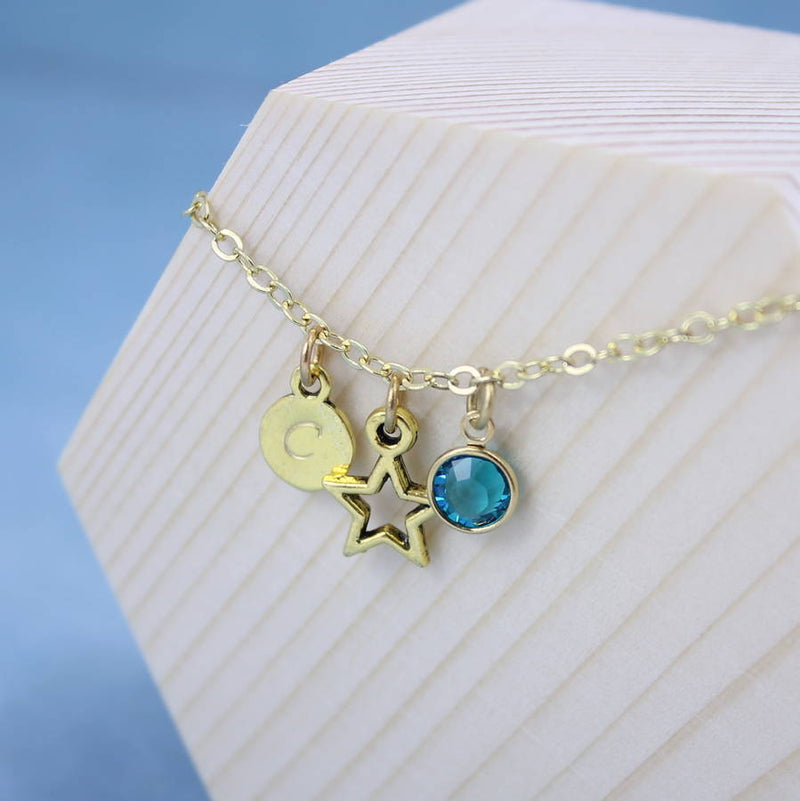 Image shows personalised gold star birthstone charm bracelet with C initial on disc and September birthstone,hanging on a wooden block