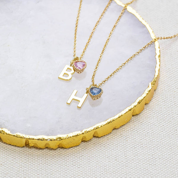 Two gold necklaces display on a jewellery dish, one necklace is a Light Amethyst heart birthstone and gold B initial charm and the 2nd necklace  has a September heart birthstone and a H initial  gold charm