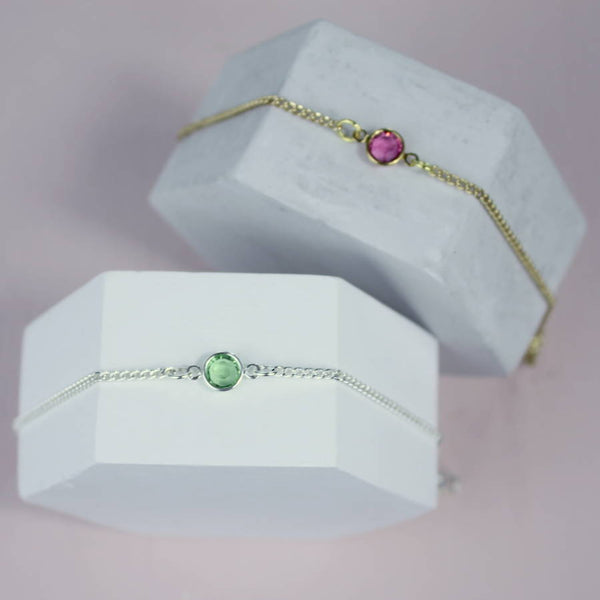 image shows personalised skinny birthstone bracelet in gold with a Rose - October birthstone and in silver with a peridot - August birthstone displayed on white hexagon blocks