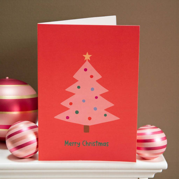 Pink Christmas tree illustration on a red greeting card with colourful baubles, a small star on top, and the text ‘Merry Christmas’ beneath