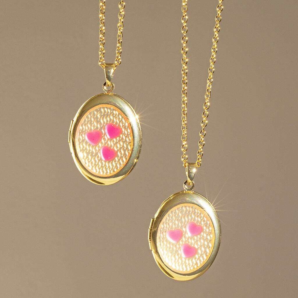 Image shows two Pink Hearts Gold Plated Oval Locket Necklaces hanging in front of a nude backdrop.