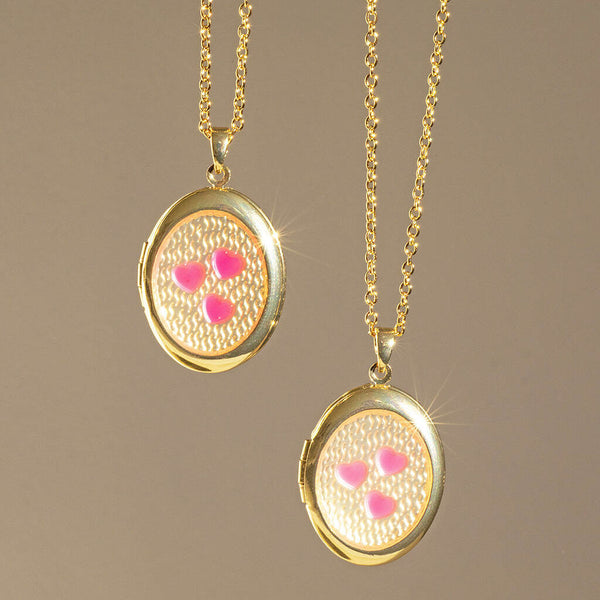 Image shows two Pink Hearts Gold Plated Oval Locket Necklaces hanging in front of a nude backdrop.