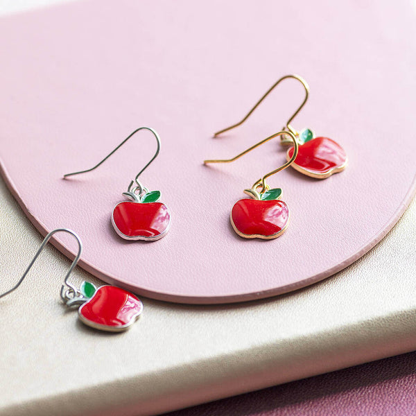 Red enamel apple charms on silver and gold earring hooks laid flat on a pink plate.