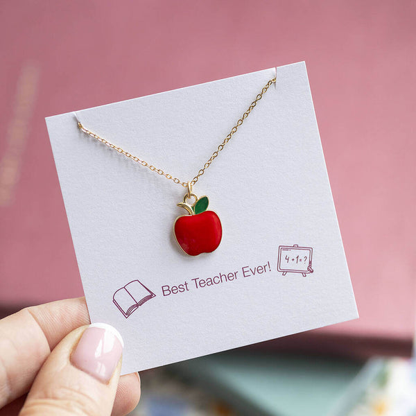 Gold necklace with a red enamel apple charm suspended on chain and presented on a Best Teacher Ever sentiment card. Pink and floral journals are in the background.