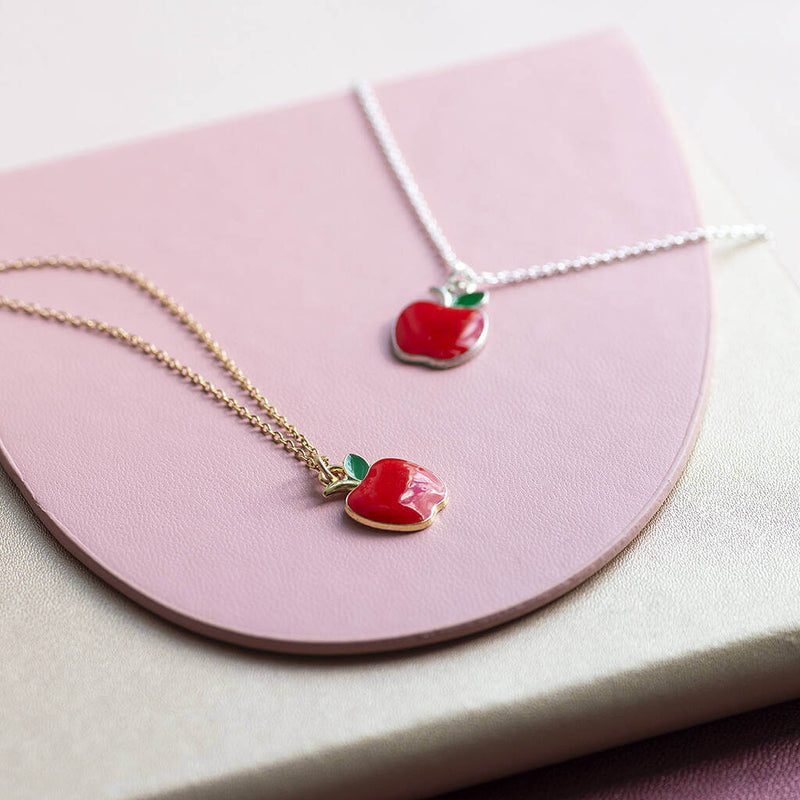Gold and silver apple charm necklaces lying flat on a pink plate onto of a cream leather jotter