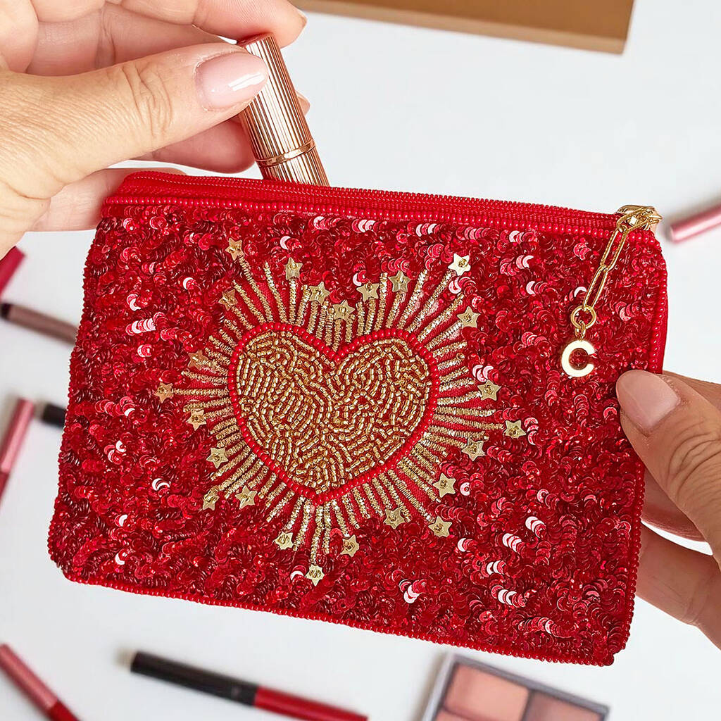 Close-up of red pouch with intricate gold beadwork in heart shape and a gold initial charm on the zip pull.