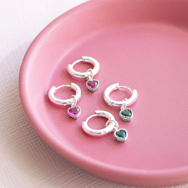 Silver plated huggie hoop earrings displayed on a pink jewellery dish. First pair with the Ruby July birthstone hearts and 2nd pair with the emerald May birthstone hearts