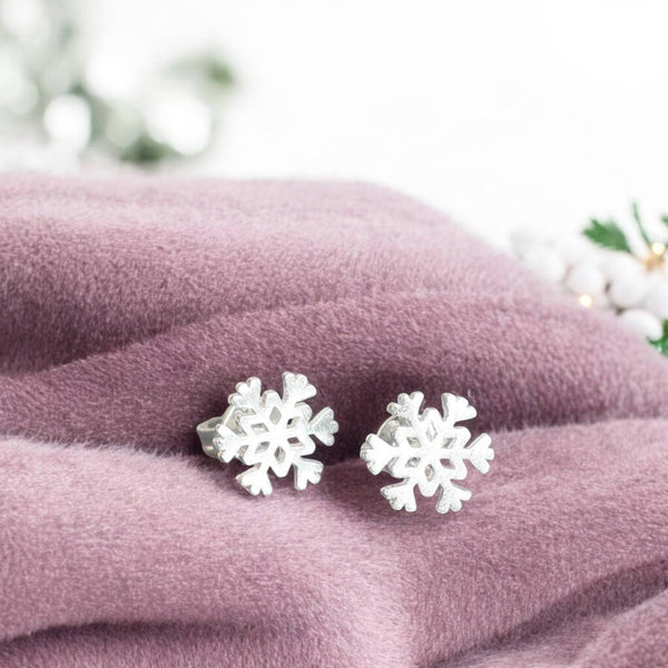 Image shows silver plated snowflake charm stud earrings presented on pink fabric with fairy lights in the background.