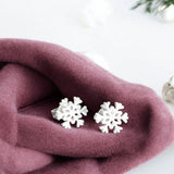 Image shows silver plated snowflake charm stud earrings presented on pink fabric with fairy lights in the background.