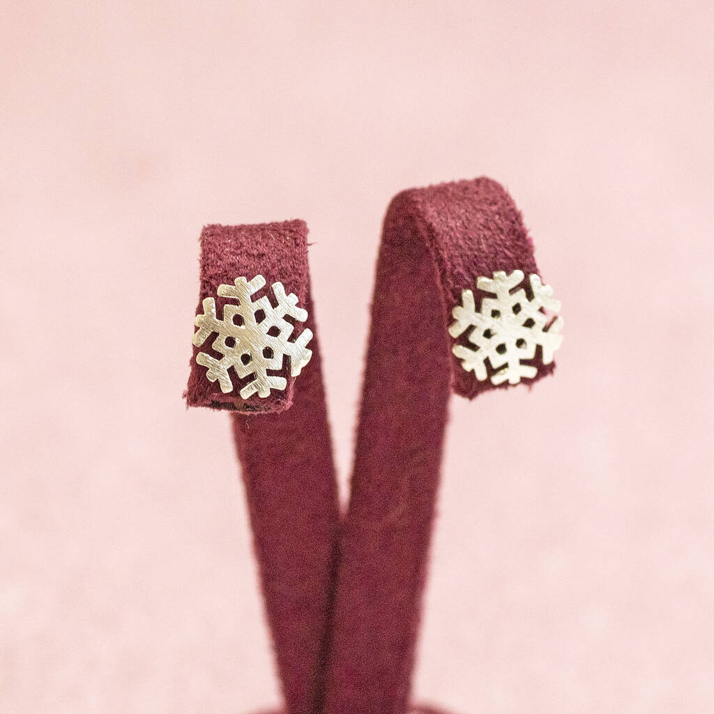 Image shows silver plated snowflake charm stud earrings presented on pink fabric jewellery stand.