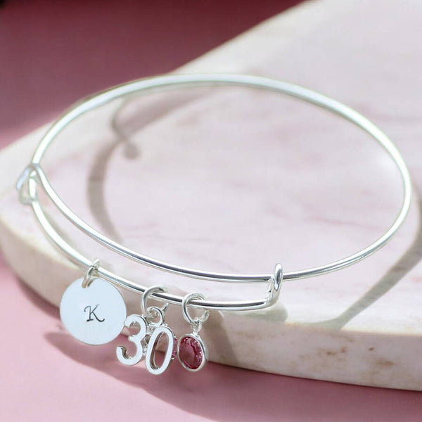 Silver personalised bangle with letter charm, 30 pendant and pink crystal birthstone on marble